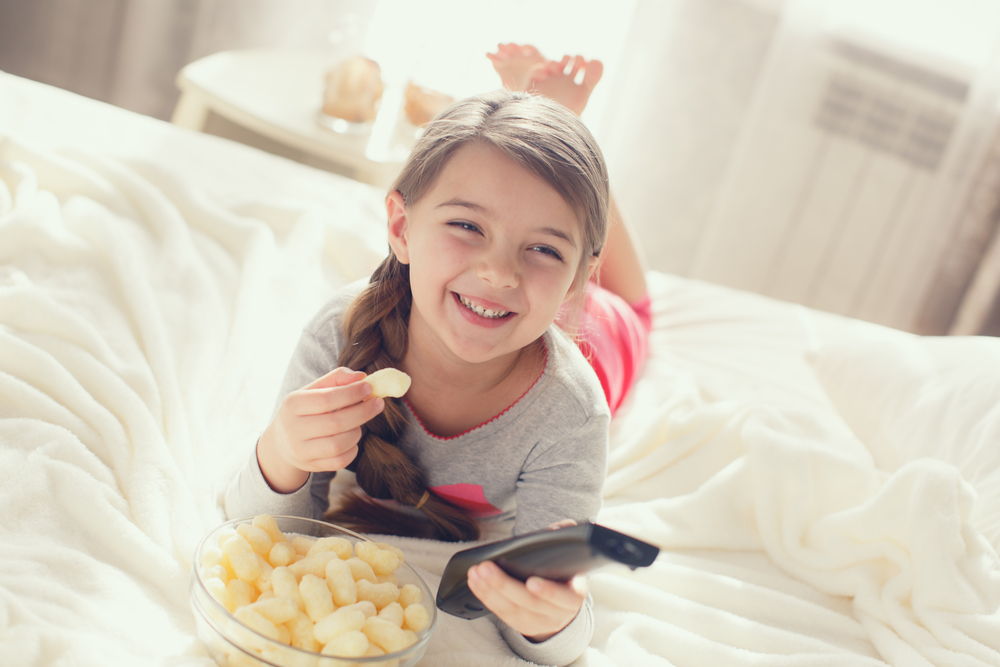 Девочка лежит на кровати с попкорном и пультом